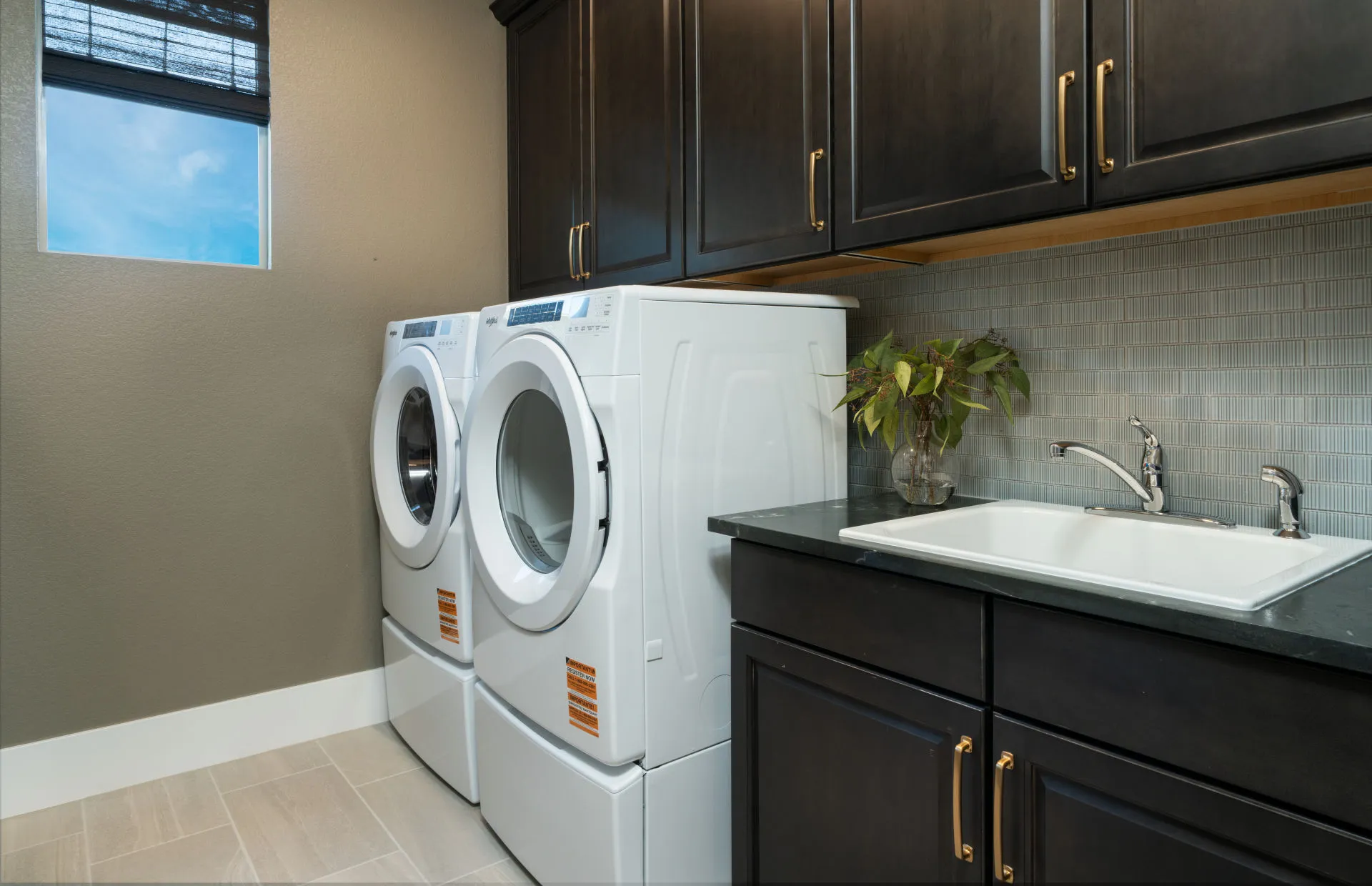 Brantley-Tivoli-Laundry Room Modern laundry room in Tivoli at Brantley by Pulte Homes features a front-loading washer and dryer, dark cabinets with gold handles, a white utility sink, tile flooring, a small potted plant, and natural light from the window.
