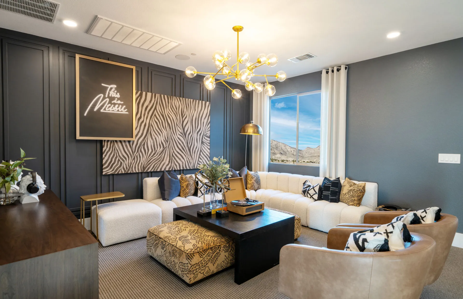 Brantley-Tivoli-Loft Modern living room at Tivoli at Brantley by Pulte Homes with a dark accent wall, gold chandelier, large beige sectional sofa, patterned pillows, black coffee table, zebra-print artwork, mountain view window, plants, and stylish accessories.