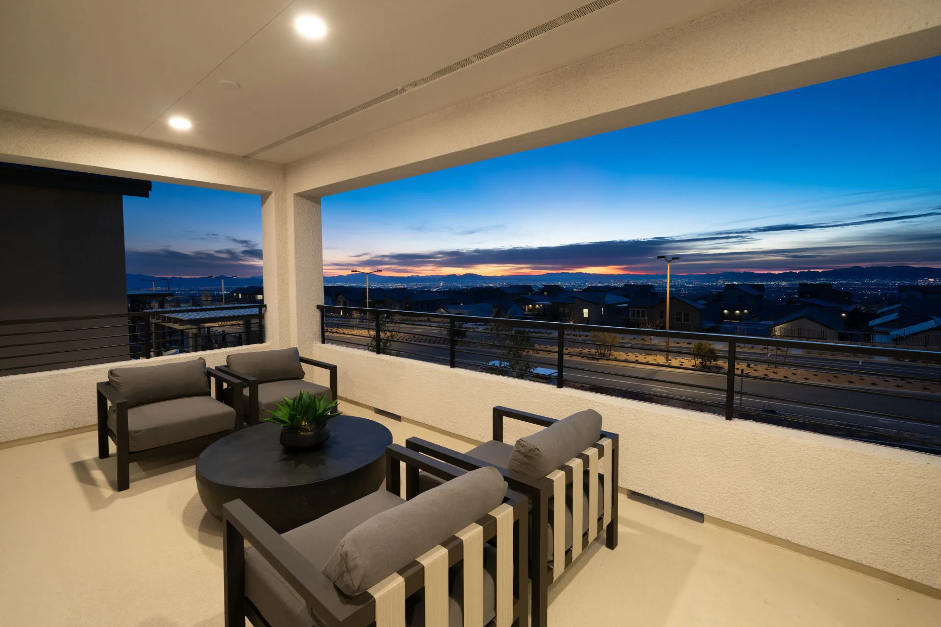 Brantley-Tivoli-Mirador Modern covered balcony at Tivoli at Brantley by Pulte Homes, with cushioned chairs and a round table, overlooking a suburban neighborhood at sunset with city lights and mountains in the distance.