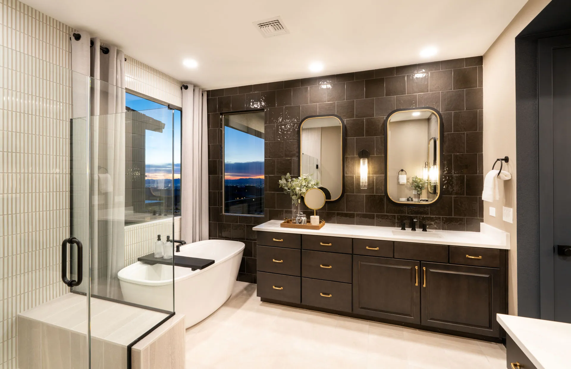Brantley-Tivoli-Owner’s Bath Modern bathroom at Tivoli at Brantley by Pulte Homes features a freestanding tub by a large window, glass shower enclosure, double vanity with dark cabinets, two mirrors, and decorative lighting; cityscape glows at sunset.