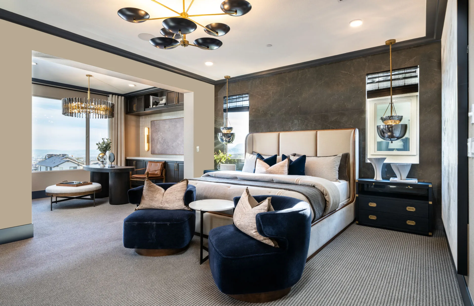 Brantley-Tivoli-Owner’s Suite Modern, spacious bedroom at Tivoli at Brantley by Pulte Homes with a neutral palette, large bed, two blue velvet chairs, a round table, work desk by the window, and striking statement light fixtures from the ceiling.