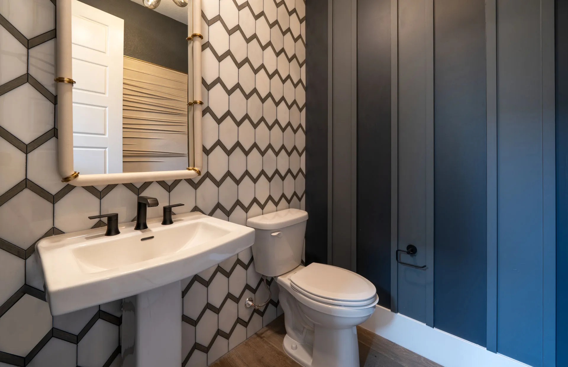 Brantley-Tivoli-Powder Room A modern bathroom at Tivoli at Brantley by Pulte Homes features a pedestal sink, black fixtures, a toilet, wood-look flooring, a hexagon-tiled wall, a rectangular mirror, and a navy blue vertical-paneled accent wall.