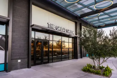 Exterior view of The Great Greek restaurant with large glass windows, a modern facade, and an overhang. Decorative plants are near the entrance, and the interior is partially visible through the glass.