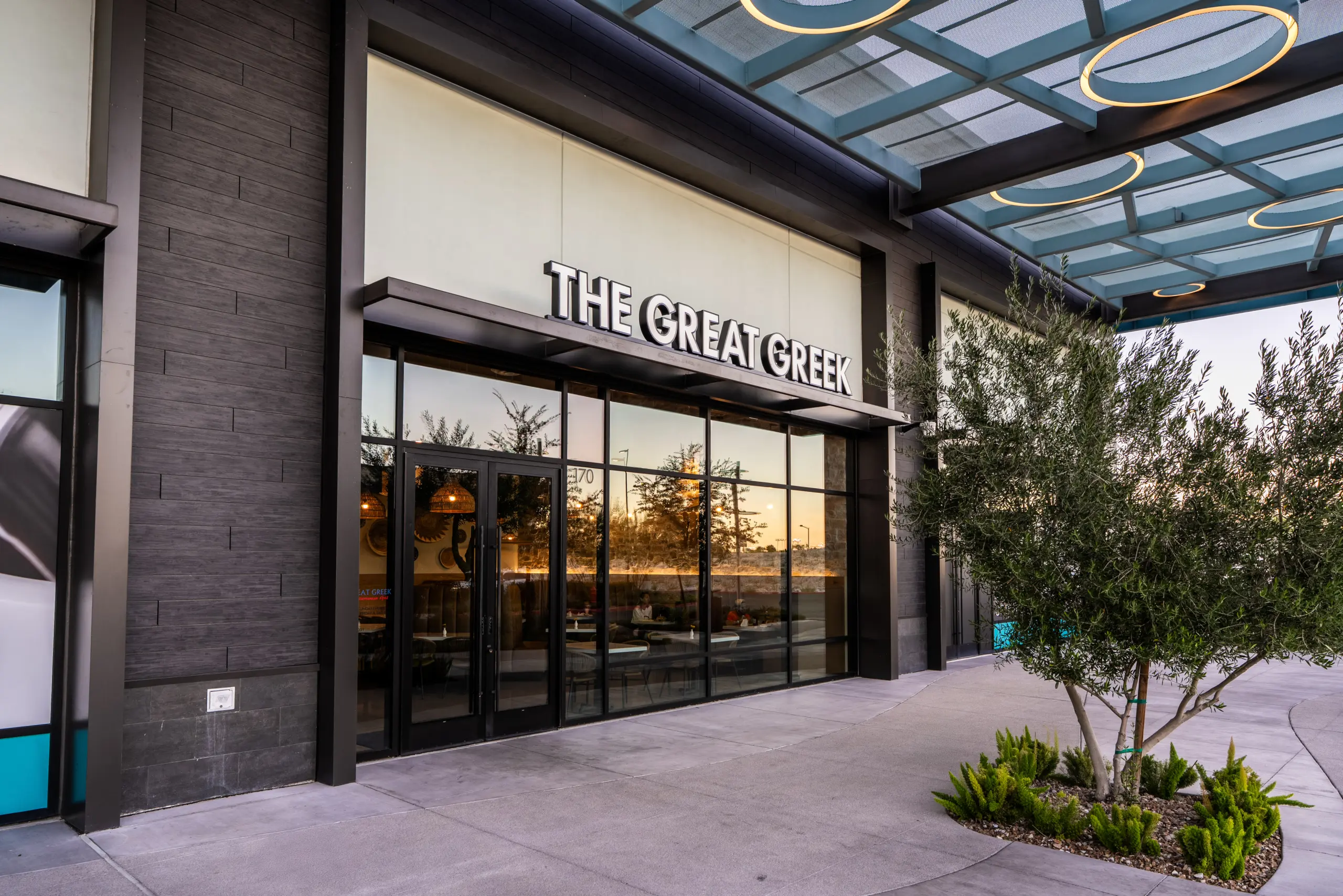 Exterior view of The Great Greek restaurant with large glass windows, a modern facade, and an overhang. Decorative plants are near the entrance, and the interior is partially visible through the glass.