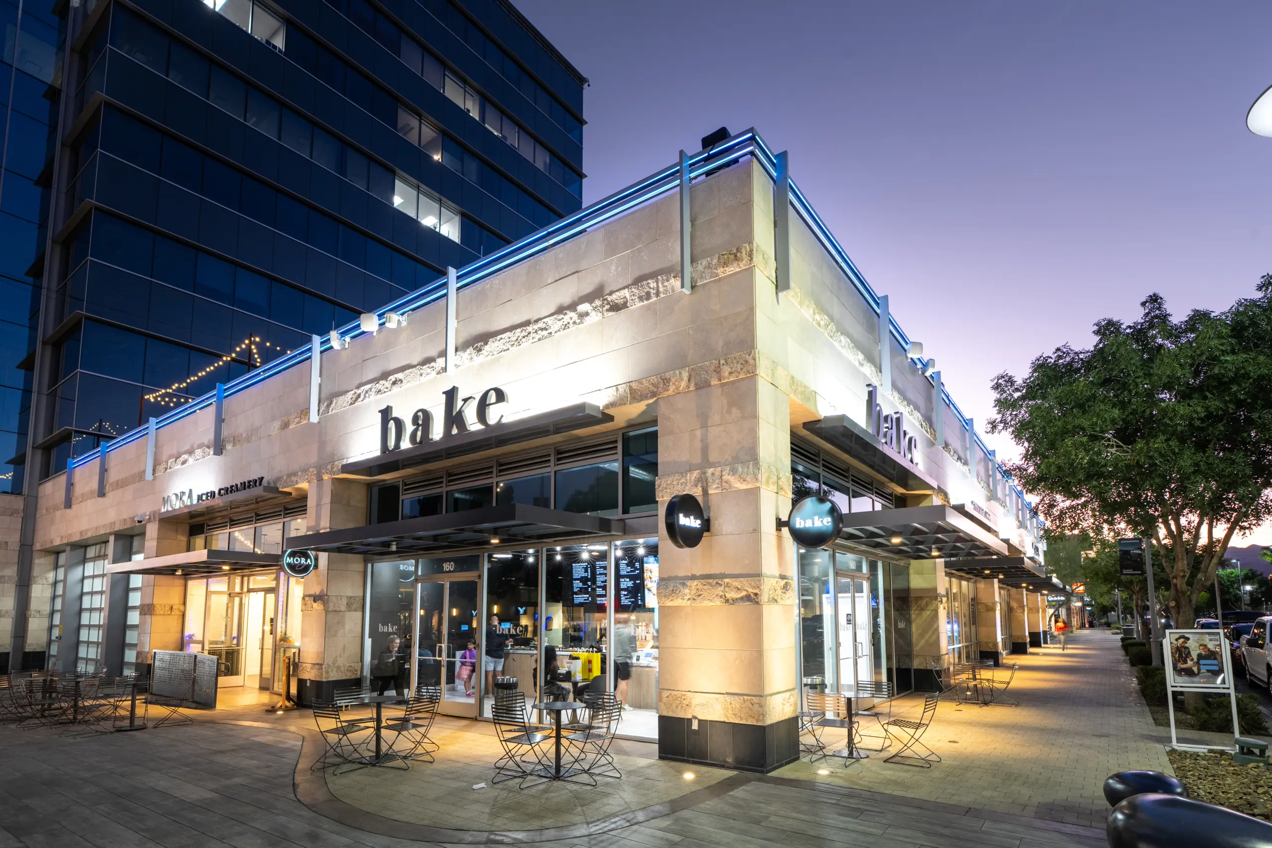 DSC06053 A modern bakery café called “bake” with large windows, illuminated signage, and outdoor seating on a city sidewalk at dusk. The sky is purple, and the building is next to a tall glass office. Trees and lights line the street.