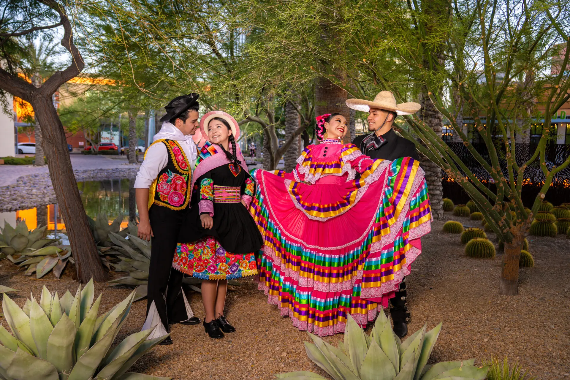 Four people in vibrant traditional Mexican folkloric attire stand outdoors among succulents and trees, smiling and posing. Two women wear colorful dresses, and two men wear embroidered outfits with wide-brimmed hats.