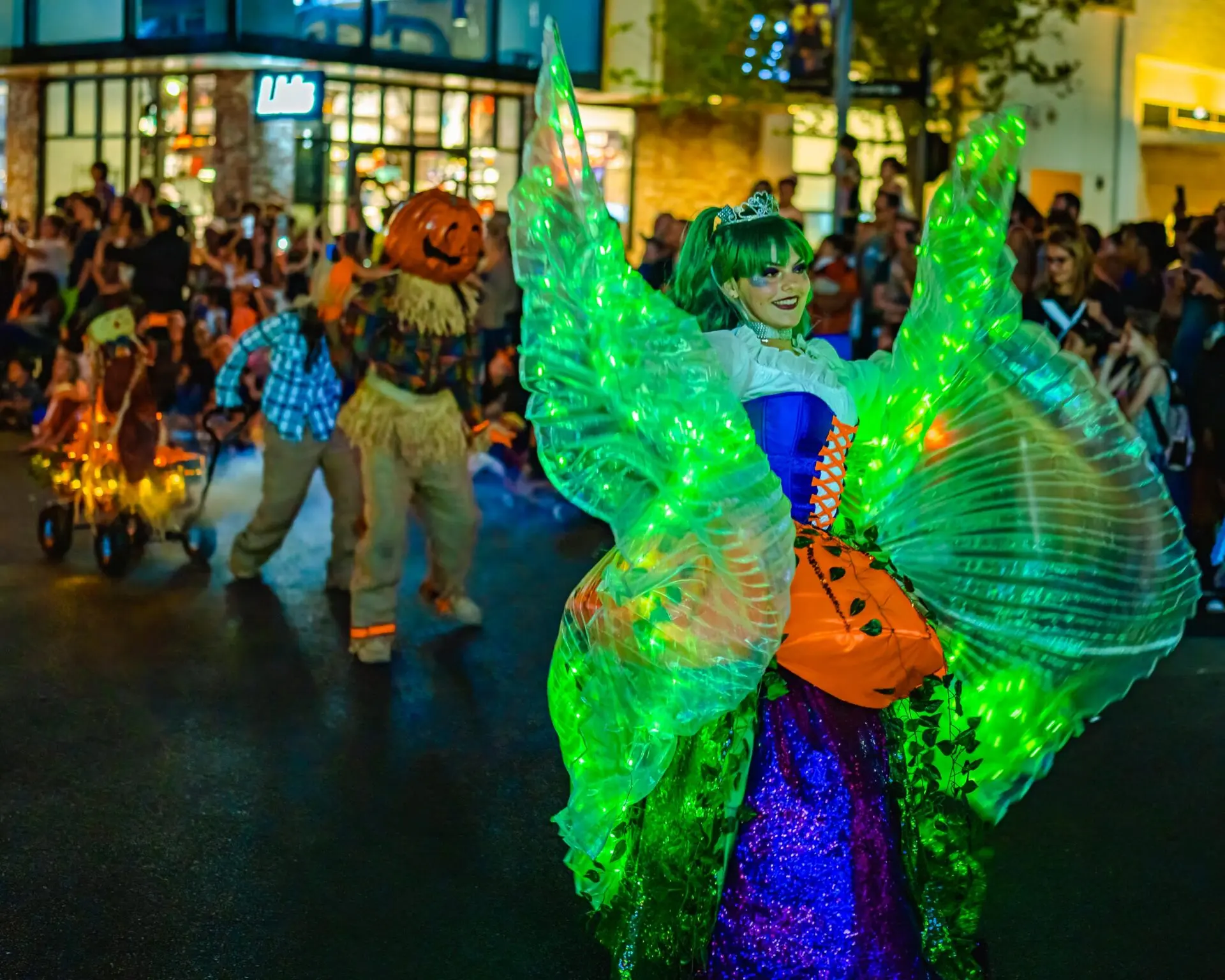 A woman in a colorful, glowing costume with large green illuminated wings dances in a nighttime parade, while a person in a scarecrow outfit and a decorated bicycle follow behind, with a crowd watching in the background.