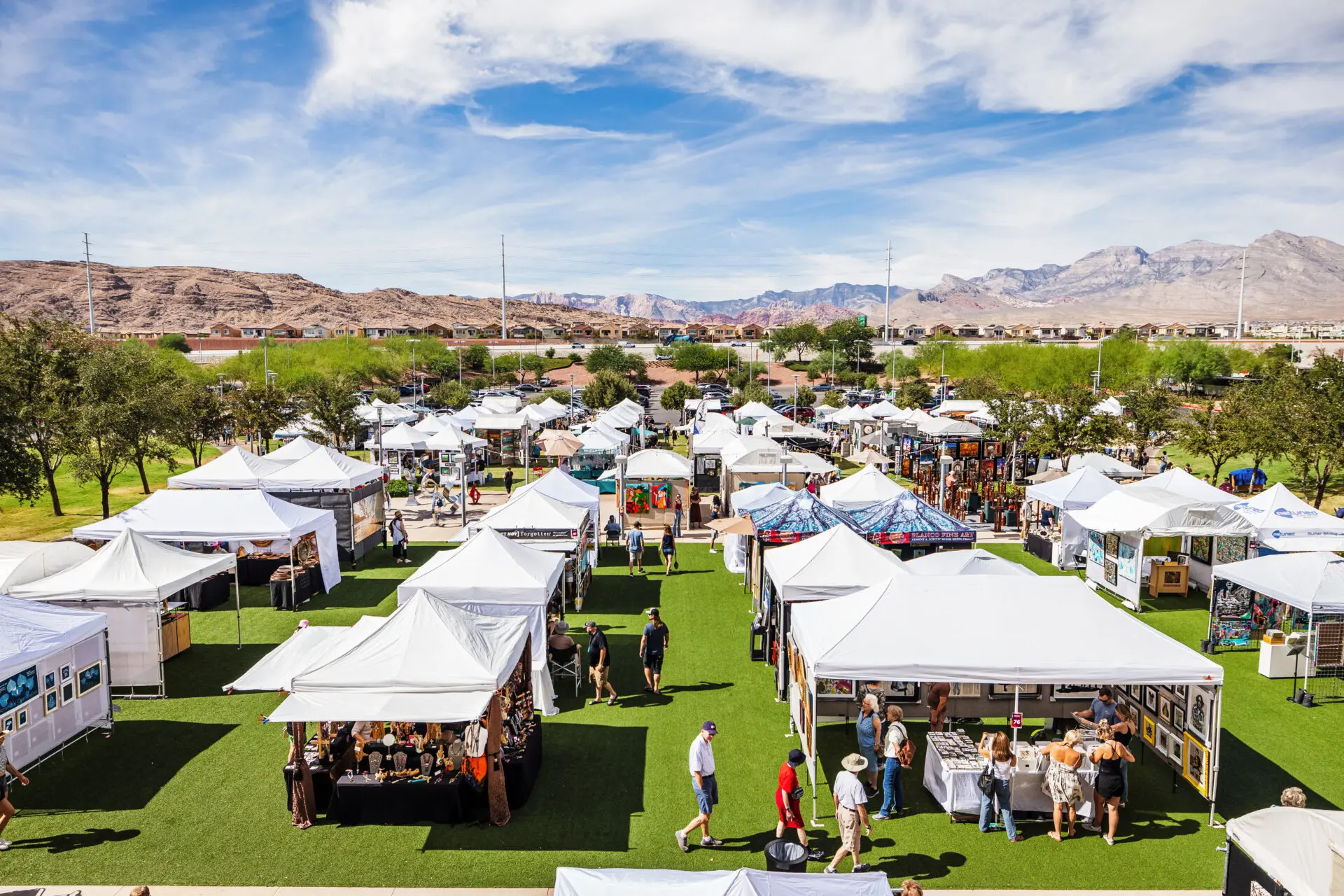 A vibrant outdoor art festival features rows of white tents on green grass, with people browsing artwork and crafts. Mountains and a blue sky with clouds are visible in the background.