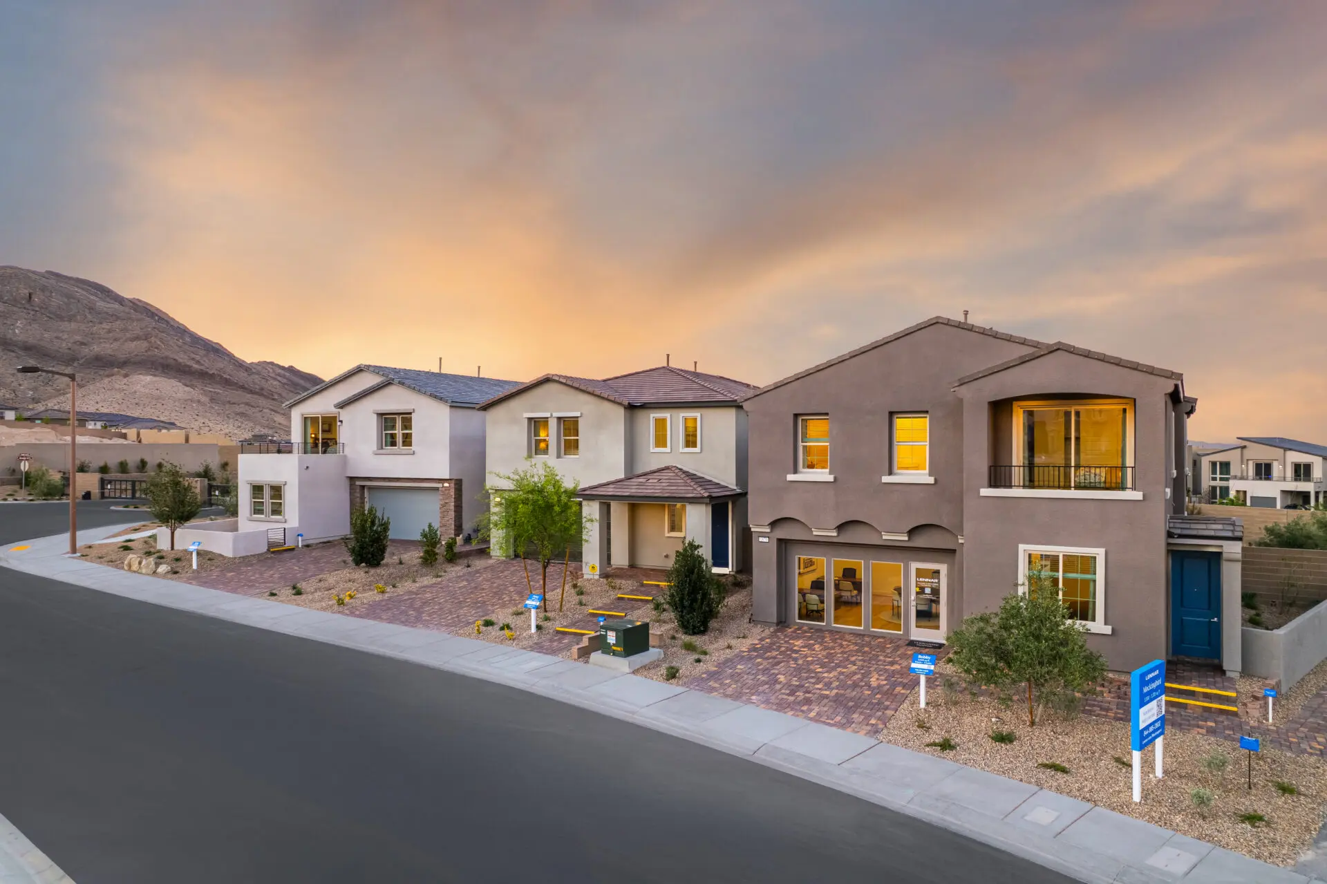 Summerlin Mockingbird by Lennar-1 A row of modern, two-story houses with landscaped yards sits on a sloped street at sunset. Warm lights glow from the windows, and desert mountains are visible in the background.