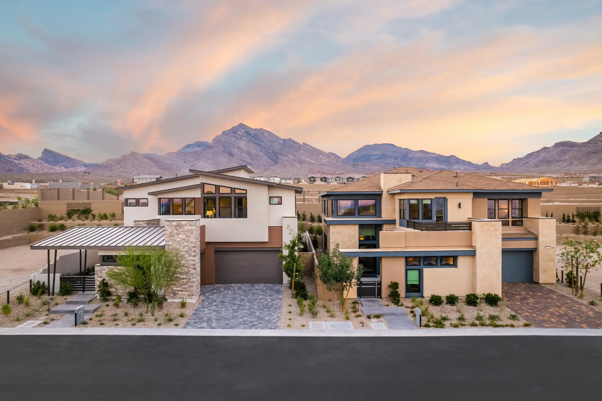 Two modern, multi-story houses with flat roofs and large windows sit side by side, surrounded by desert landscaping, with mountains and a colorful sunset sky in the background.