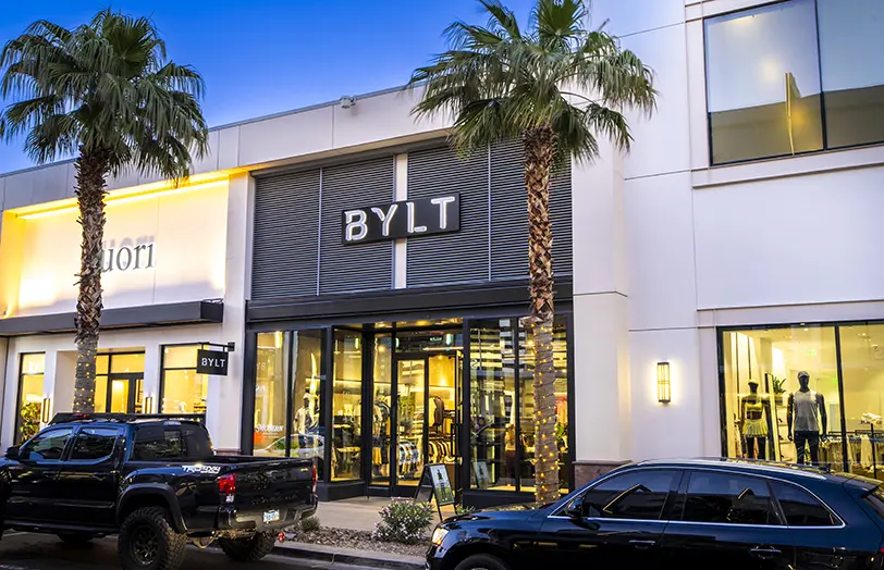 Storefront of BYLT apparel with large glass windows, palm trees, and two parked cars in front; the store interior with clothing displays is visible through the windows.