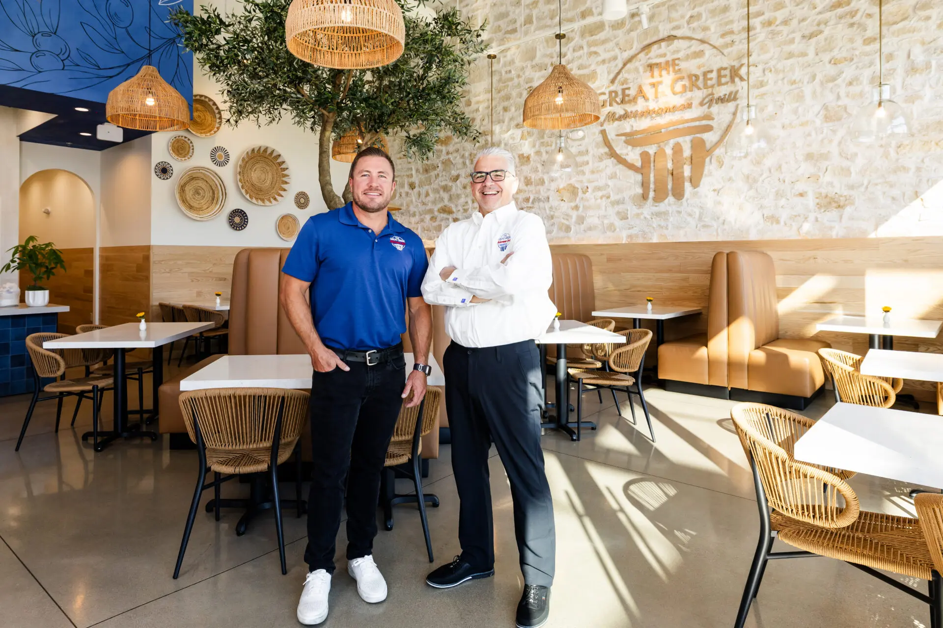 Two men stand smiling inside a bright, modern Mediterranean restaurant with wicker chairs, booth seating, wall art, and hanging lights. The sign on the wall reads The Great Greek Mediterranean Grill.