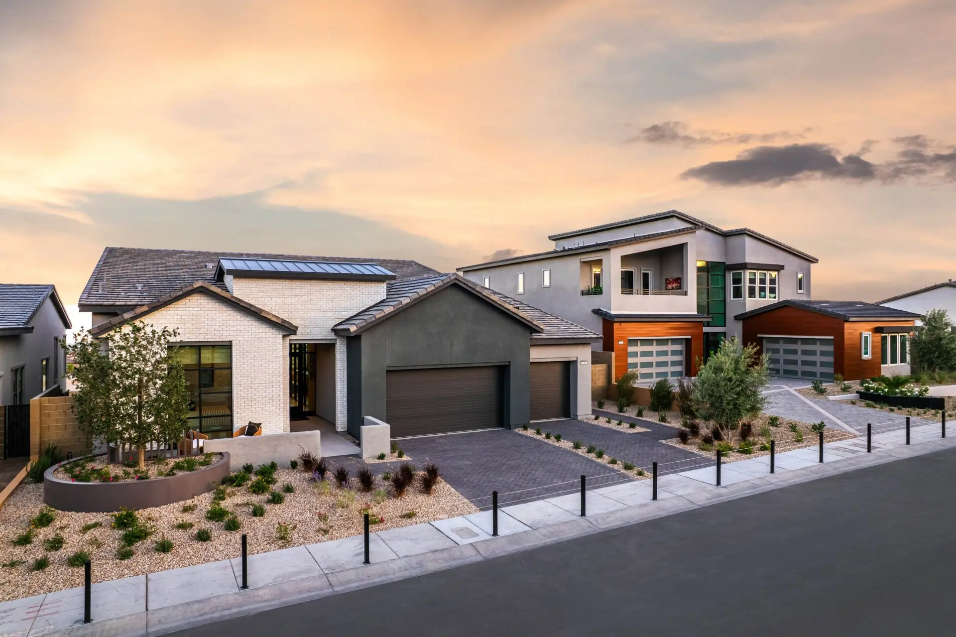 Modern suburban houses with clean lines, large windows, and landscaped front yards featuring gravel, desert plants, and small trees, set against a sunset sky on a quiet residential street.