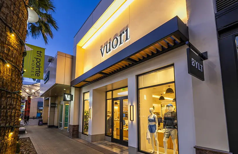 A Vuori store at dusk, with illuminated signage above glass doors and windows displaying mannequins in athletic wear; palm trees and other shop signs are visible along the sidewalk.