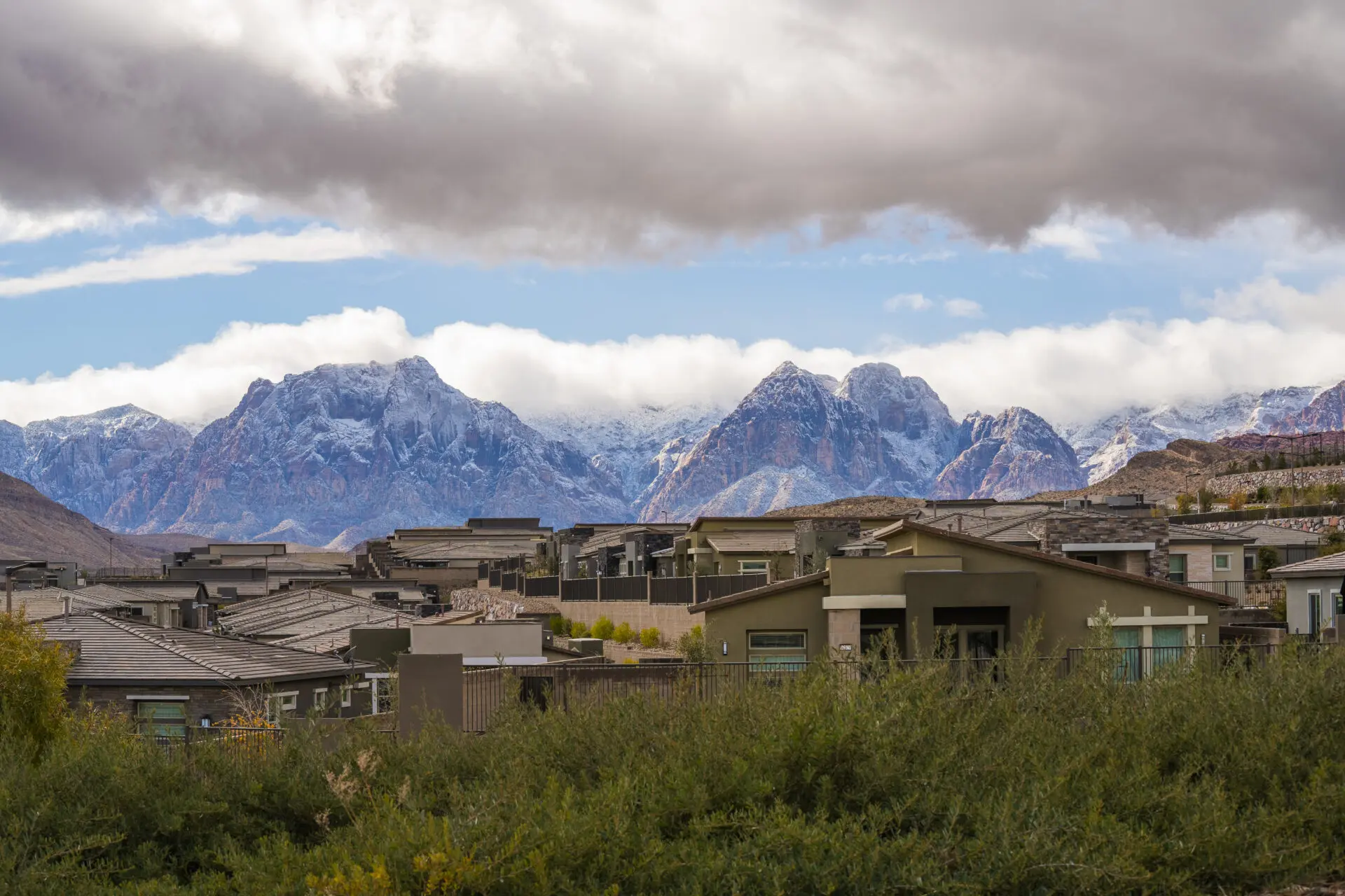 Modern houses with flat roofs are seen in the foreground, with green shrubs in front. Behind the homes, snow-capped mountains rise under a cloudy sky with patches of blue visible.