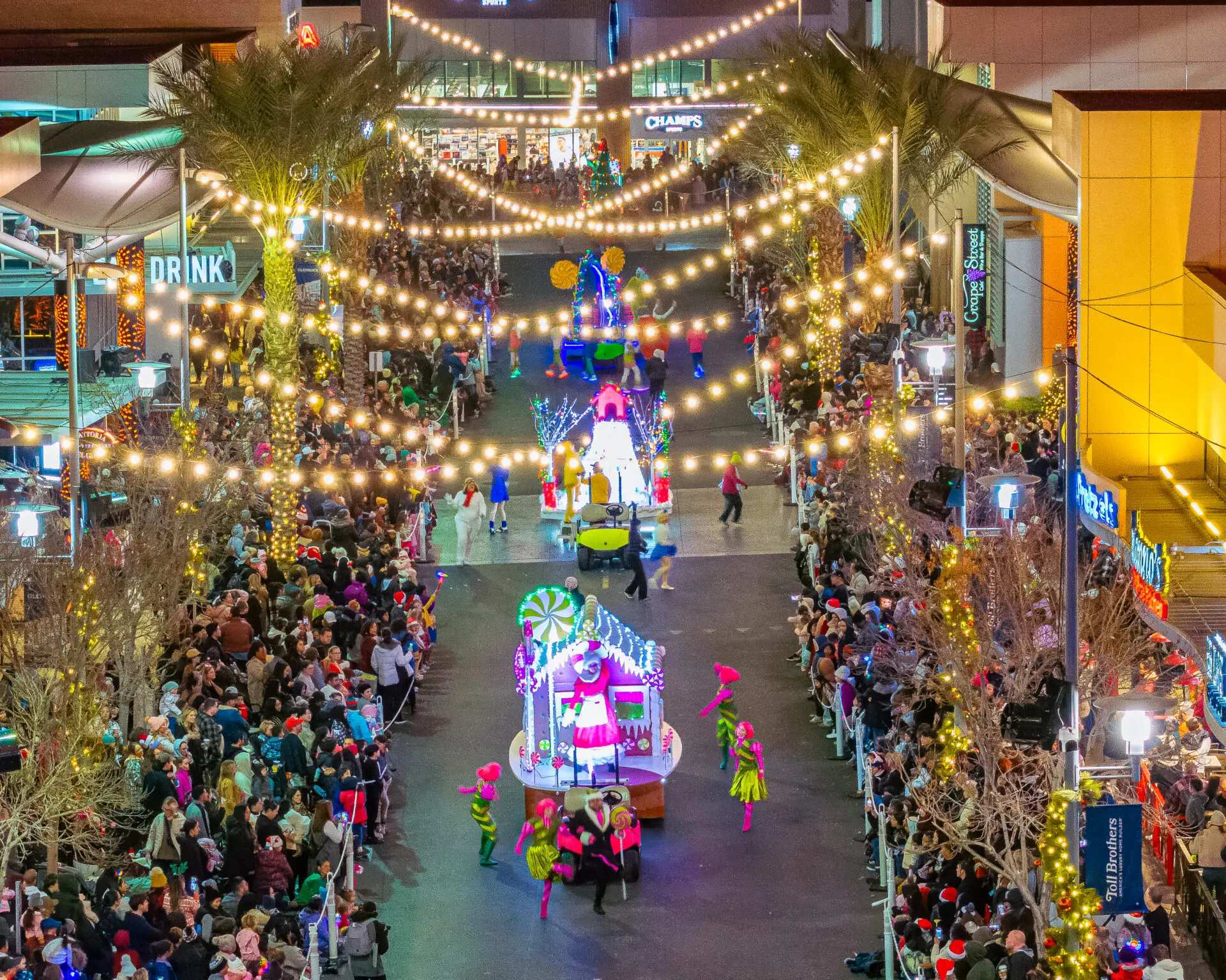 A festive parade moves down a city street lined with crowds. Colorful floats, performers in bright costumes, and overhead string lights create a lively, celebratory atmosphere at night.