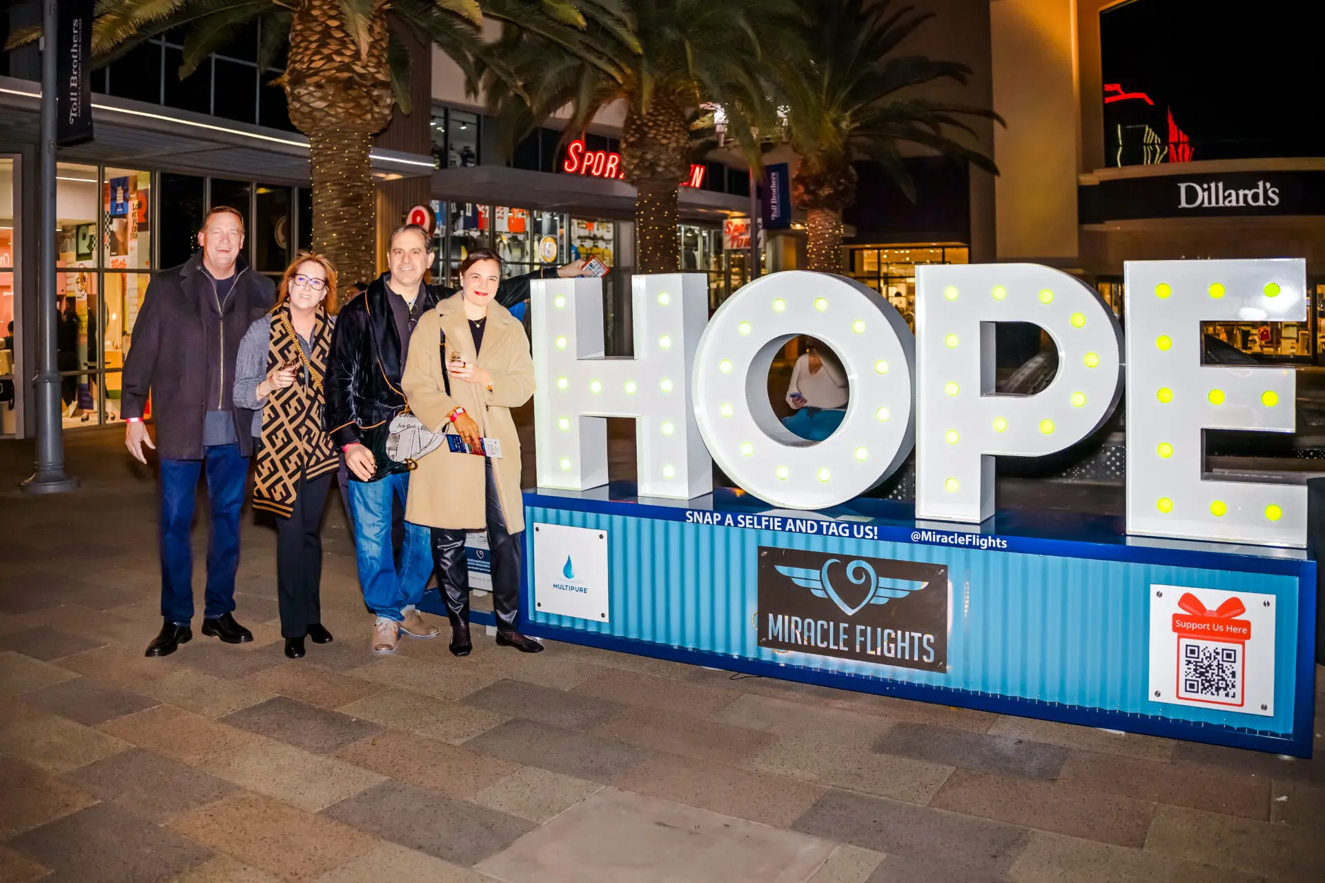 Four people stand smiling beside large, illuminated letters spelling HOPE at an outdoor shopping area at night. Signs for Miracle Flights and nearby shops are visible in the background.