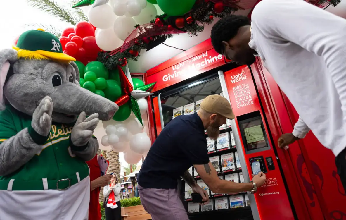 Two men use a red vending machine labeled “Giving Machine” for donations, while a person in an Oakland A’s elephant mascot costume stands nearby. Festive balloons and decorations are visible above the machine.