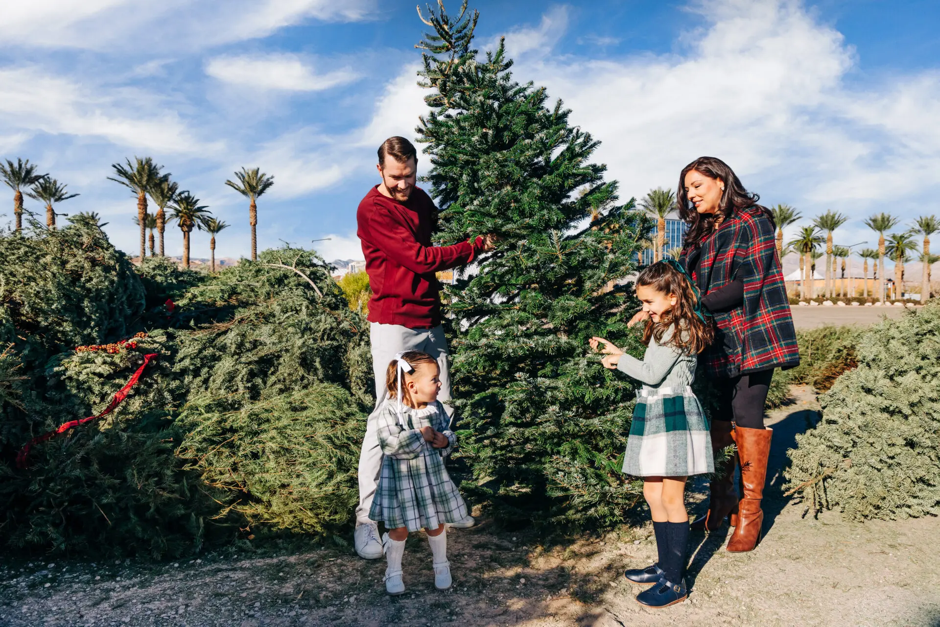 A family of four stands outdoors by a Christmas tree, smiling and decorating it together. Palm trees and blue sky are in the background on a sunny day.