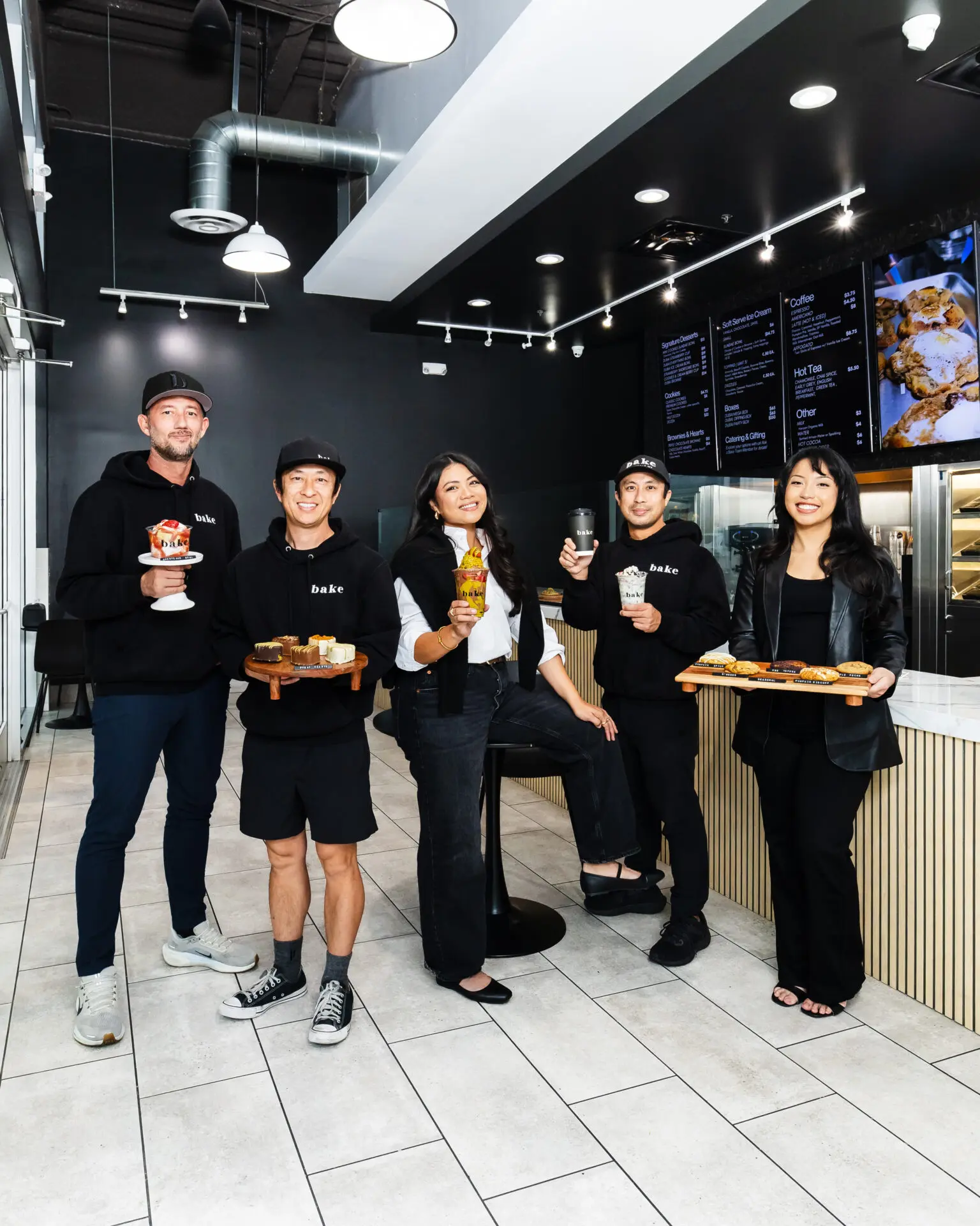 Five people stand and smile inside a modern bakery, holding various desserts and pastries. Behind them is a counter, display case with treats, and digital menu boards on the wall. The atmosphere is bright and welcoming.