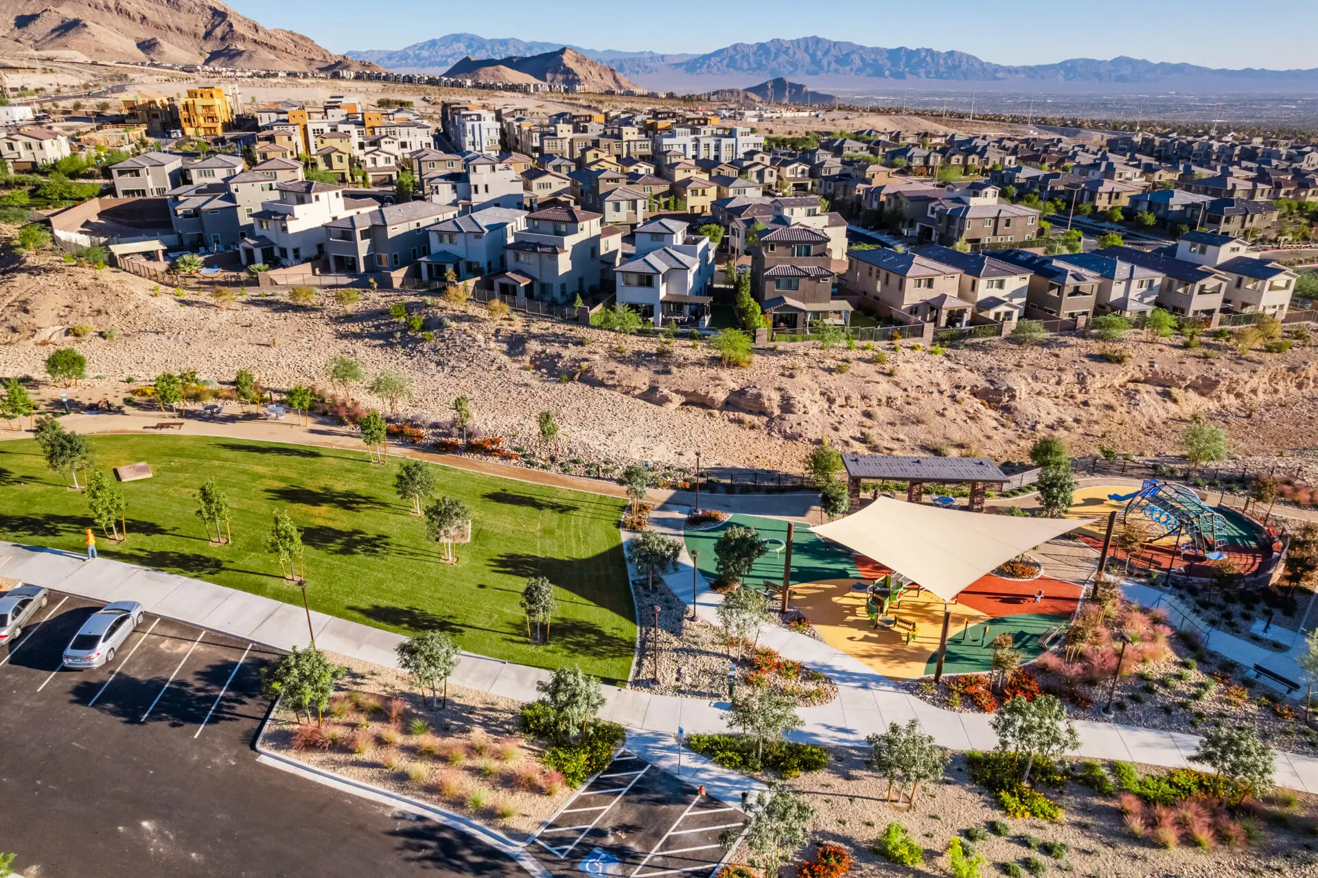 Aerial view of a suburban neighborhood with rows of modern houses, a grassy park with trees, a playground with shade structures, and mountains in the background under a clear blue sky.