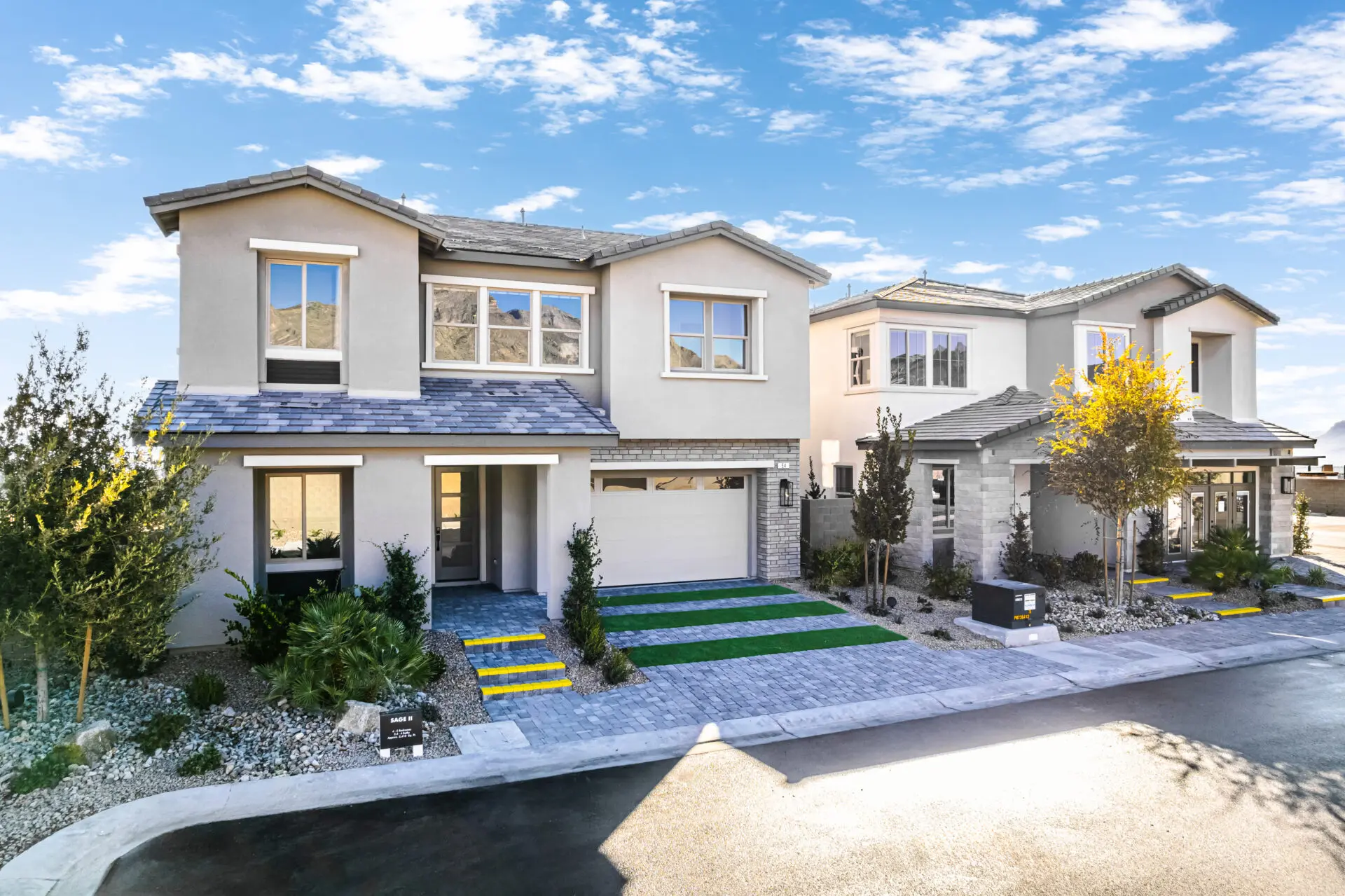 Two modern two-story houses with garages, large windows, and landscaped front yards are shown on a sunny day with a blue sky and scattered clouds. The driveways and sidewalks are accented with green and yellow stripes.