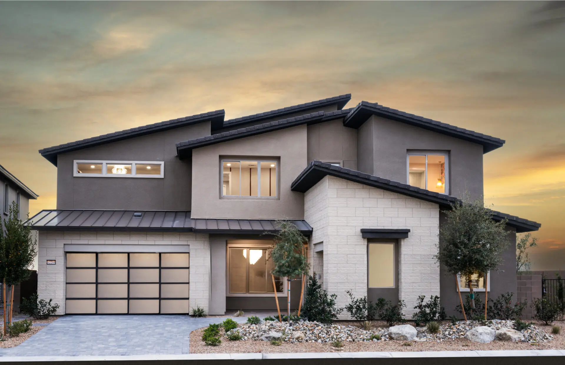 Modern two-story house with large windows, a double garage, and a mix of gray and beige exterior finishes. The front yard has small trees, rocks, and desert landscaping under a sunset sky.