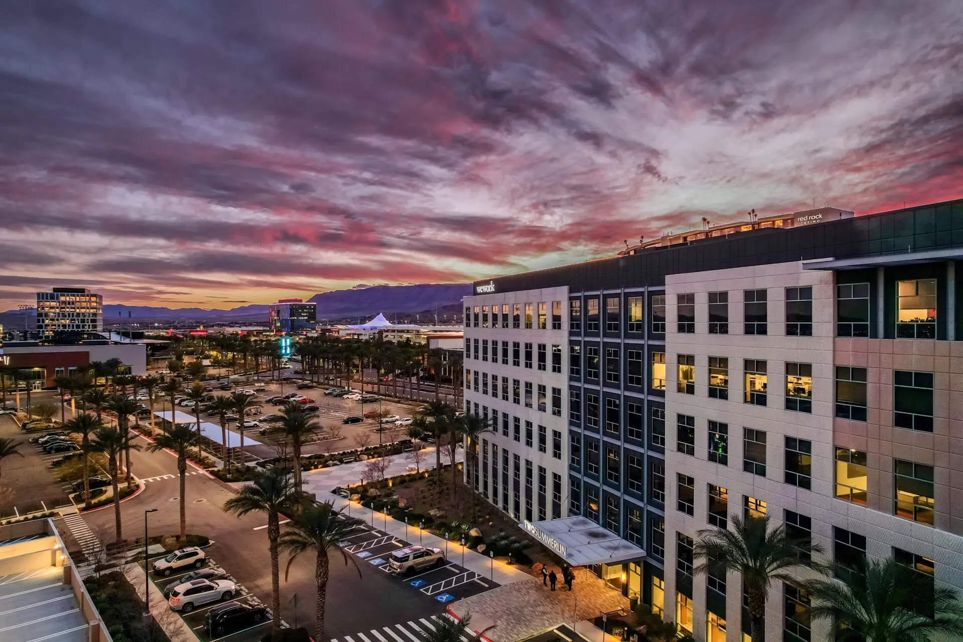 A modern office building with lit windows stands beside a palm-lined parking lot at sunset, with a dramatic, colorful sky and distant mountains in the background.