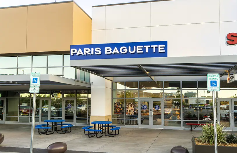Exterior of a Paris Baguette bakery with a blue sign, glass doors, blue outdoor tables and benches, and two reserved accessible parking signs in front.