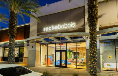 Exterior of a Roche Bobois furniture store at dusk, with a well-lit interior visible through glass windows, palm trees in front, and a car parked nearby.