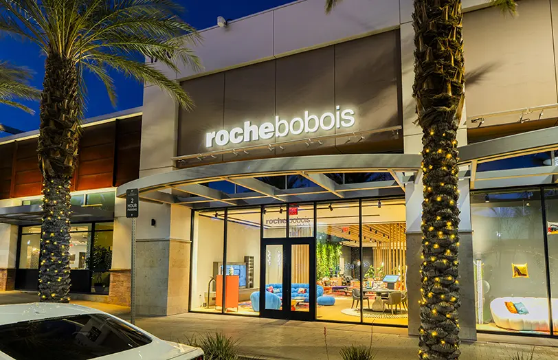 Exterior of a Roche Bobois furniture store at dusk, with a well-lit interior visible through glass windows, palm trees in front, and a car parked nearby.