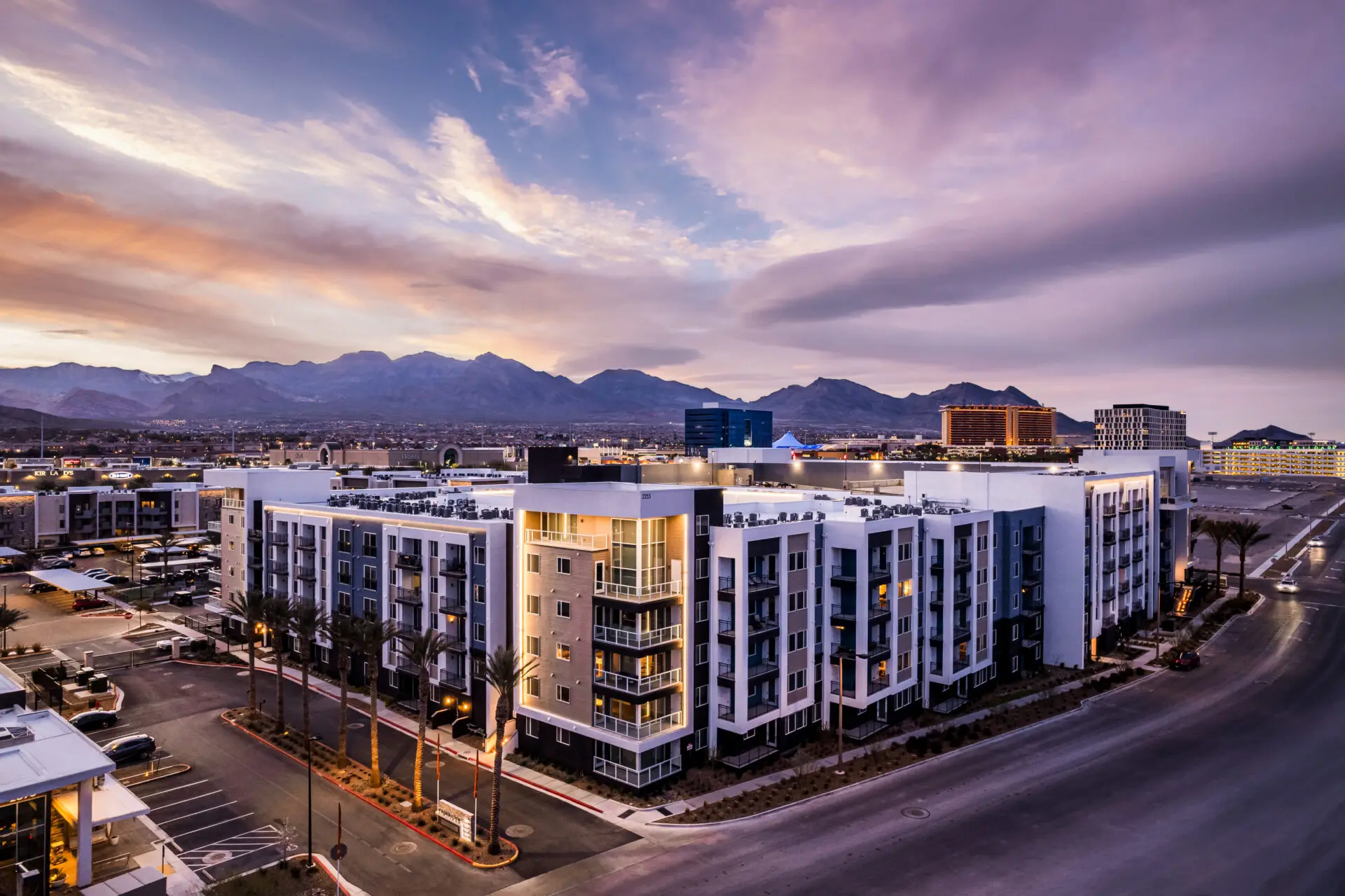 Modern apartment complex with balconies and palm trees, set against a backdrop of mountains and a dramatic sunset sky, with city buildings and roads surrounding the area.