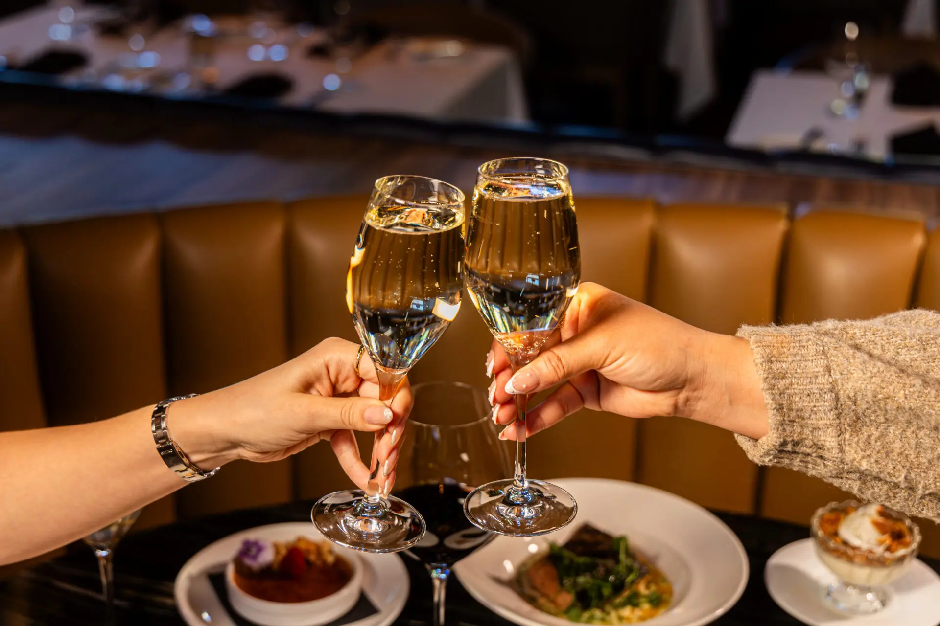 Two people clink glasses of champagne over a table set with plated dishes, including a dessert, in a warmly lit restaurant setting.