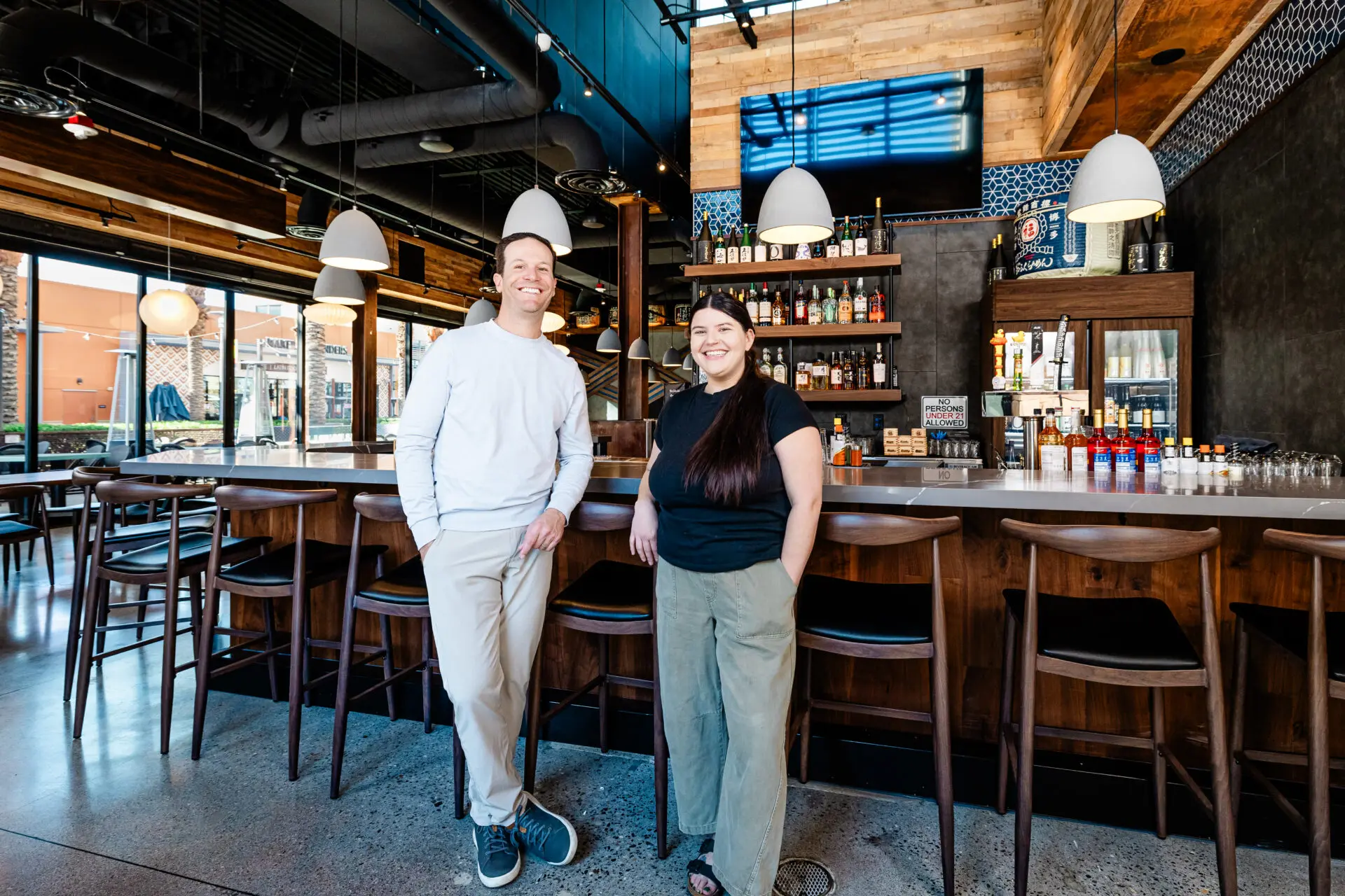 Two people stand and smile in front of a modern bar with wooden accents, pendant lights, and a well-stocked back bar. The bar stools and tables are empty, and large windows allow natural light into the space.