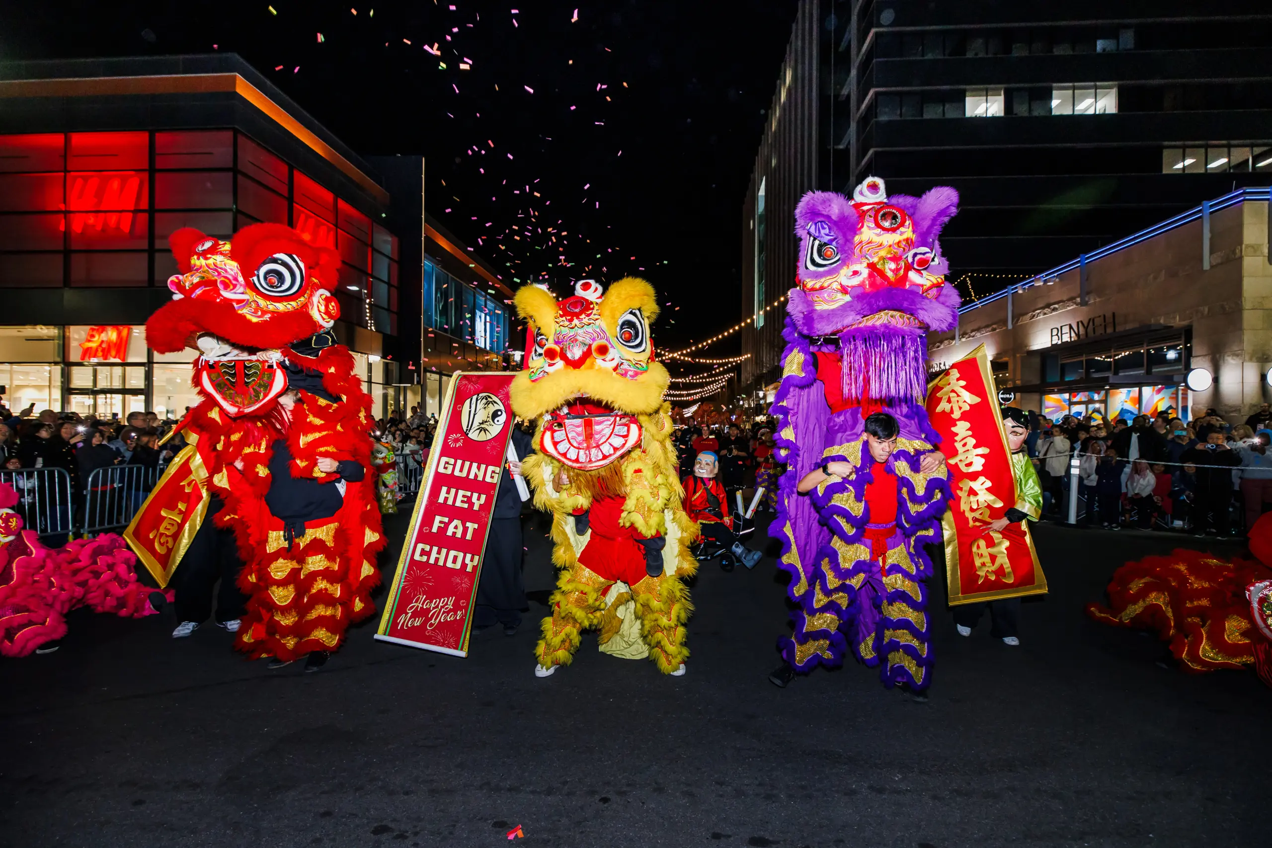 Three colorful lion dance costumes—red, yellow, and purple—perform on a city street at night during a festival, with confetti falling and a crowd watching. One lion holds a sign reading Gung Hey Fat Choy.