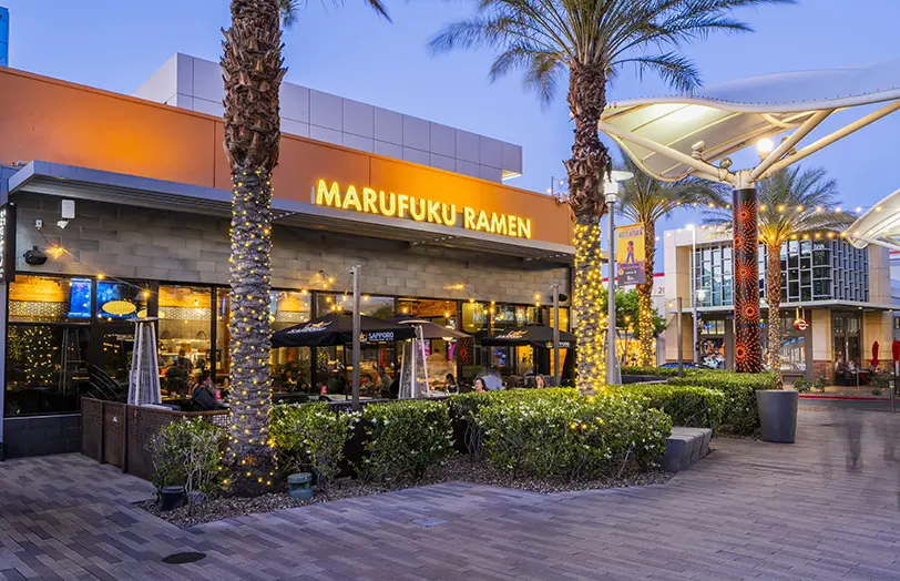 Outdoor view of Marufuku Ramen restaurant at dusk, with string lights wrapped around palm trees, patio seating, and modern architectural elements in a lively shopping area.