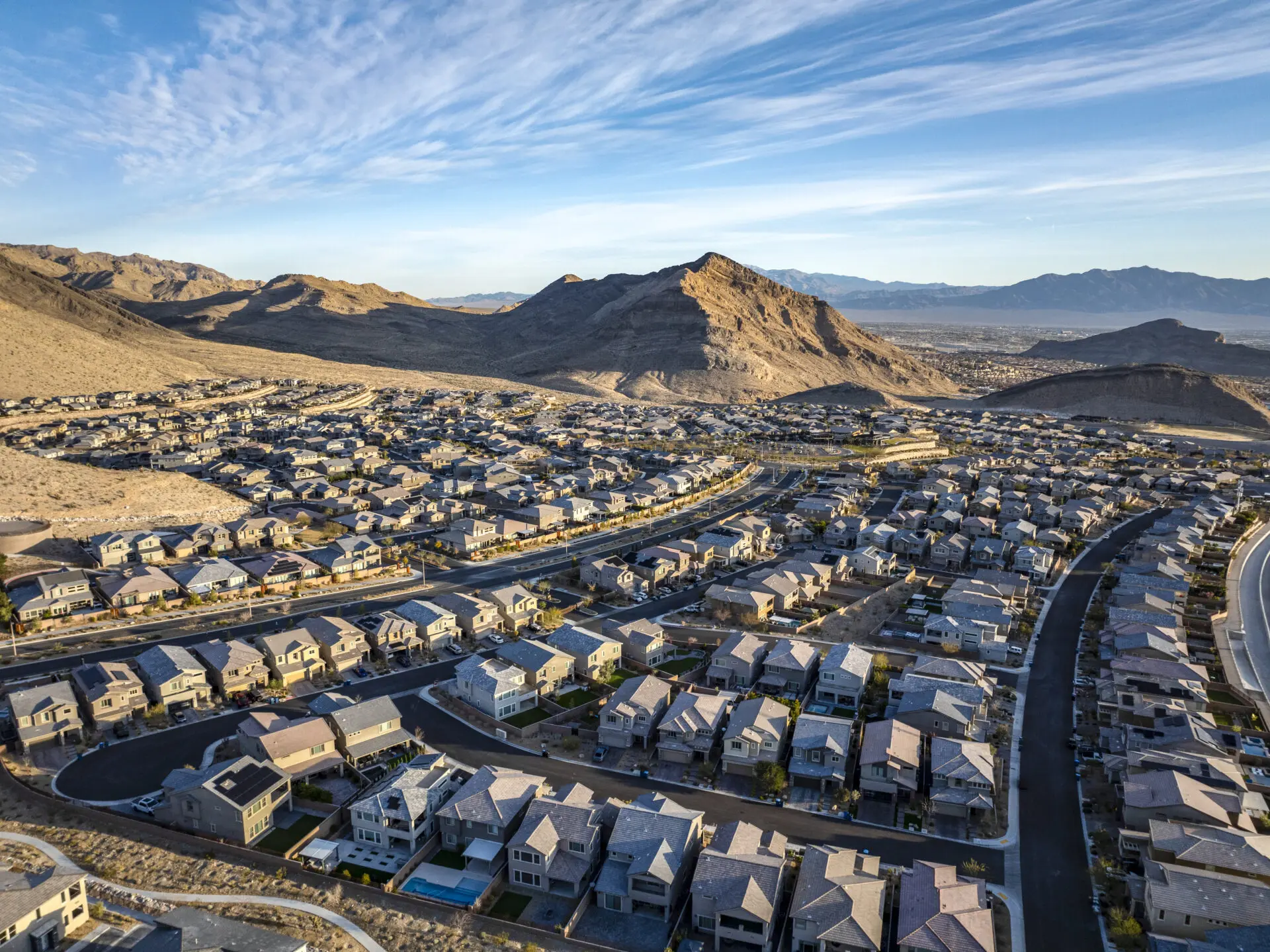 Aerial view of a suburban neighborhood with many houses, curved streets, and desert mountains in the background under a blue sky with wispy clouds.