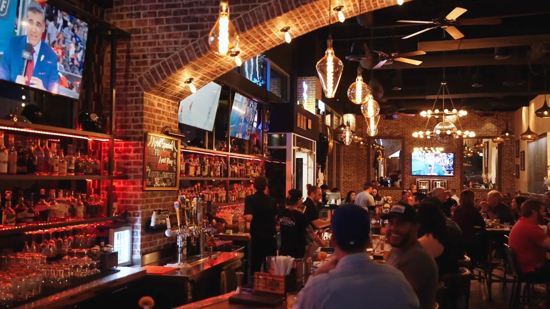 A lively bar with exposed brick walls, hanging lights, and multiple TVs showing sports. Bartenders serve drinks behind the bar while patrons sit, socialize, and watch the screens. The atmosphere is warm and bustling.