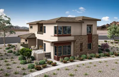 Modern two-story house with stone and stucco exterior, large windows, and a small front patio. The home is surrounded by desert landscaping with rocks, shrubs, and a few trees, set against a mountainous backdrop.