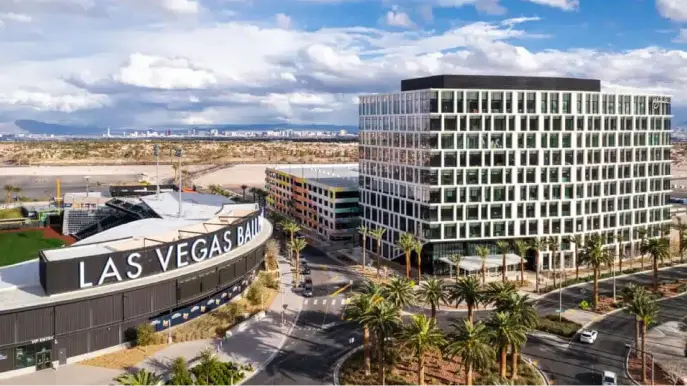 A modern city scene with the Las Vegas Ballpark in the foreground, a tall glass office building nearby, palm trees, roads, and the Las Vegas skyline visible in the distance under a partly cloudy sky.
