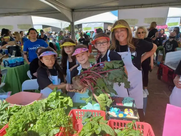 A group of children and an adult in aprons and visors smile and pose under a tent, holding a large beet. Baskets of fresh greens and vegetables are on the table in front of them at an outdoor event.
