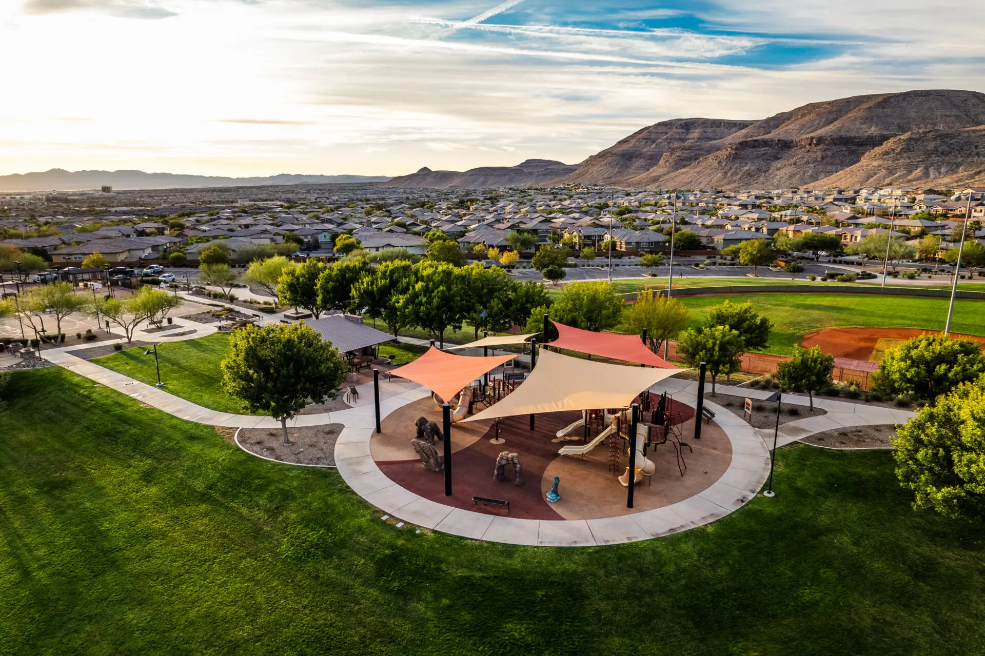 Aerial view of a park with a playground covered by colorful shade canopies, surrounded by green grass, trees, walking paths, and a baseball field, with houses and mountains in the background.