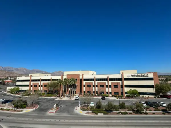 A large, modern, three-story office building with tan and red brick exterior sits under a clear blue sky, with palm trees and landscaped areas in front and mountains visible in the background.