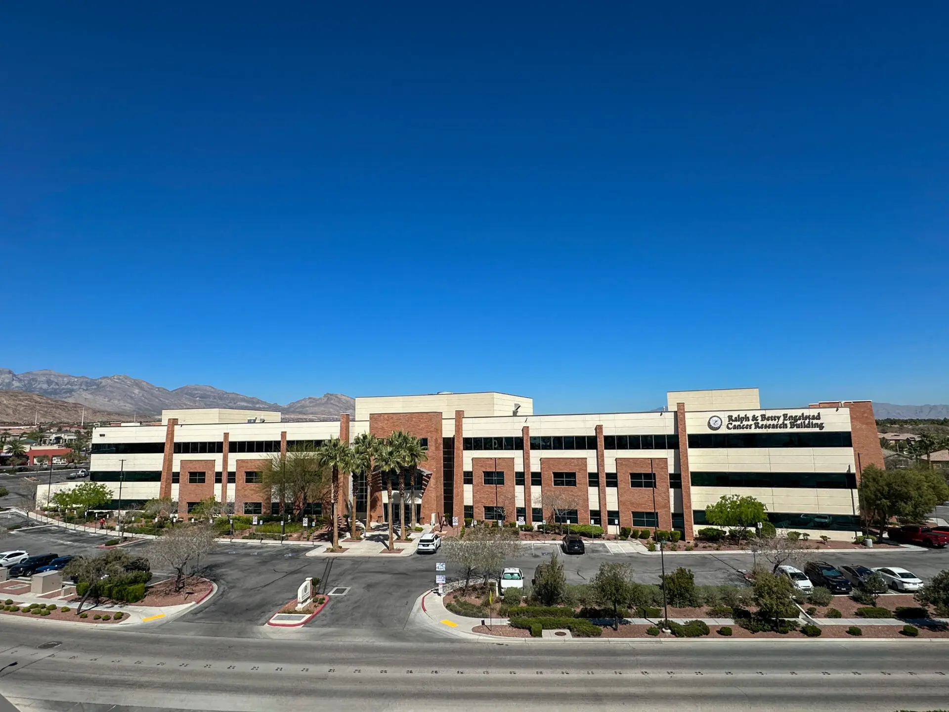 A large, modern, three-story office building with tan and red brick exterior sits under a clear blue sky, with palm trees and landscaped areas in front and mountains visible in the background.