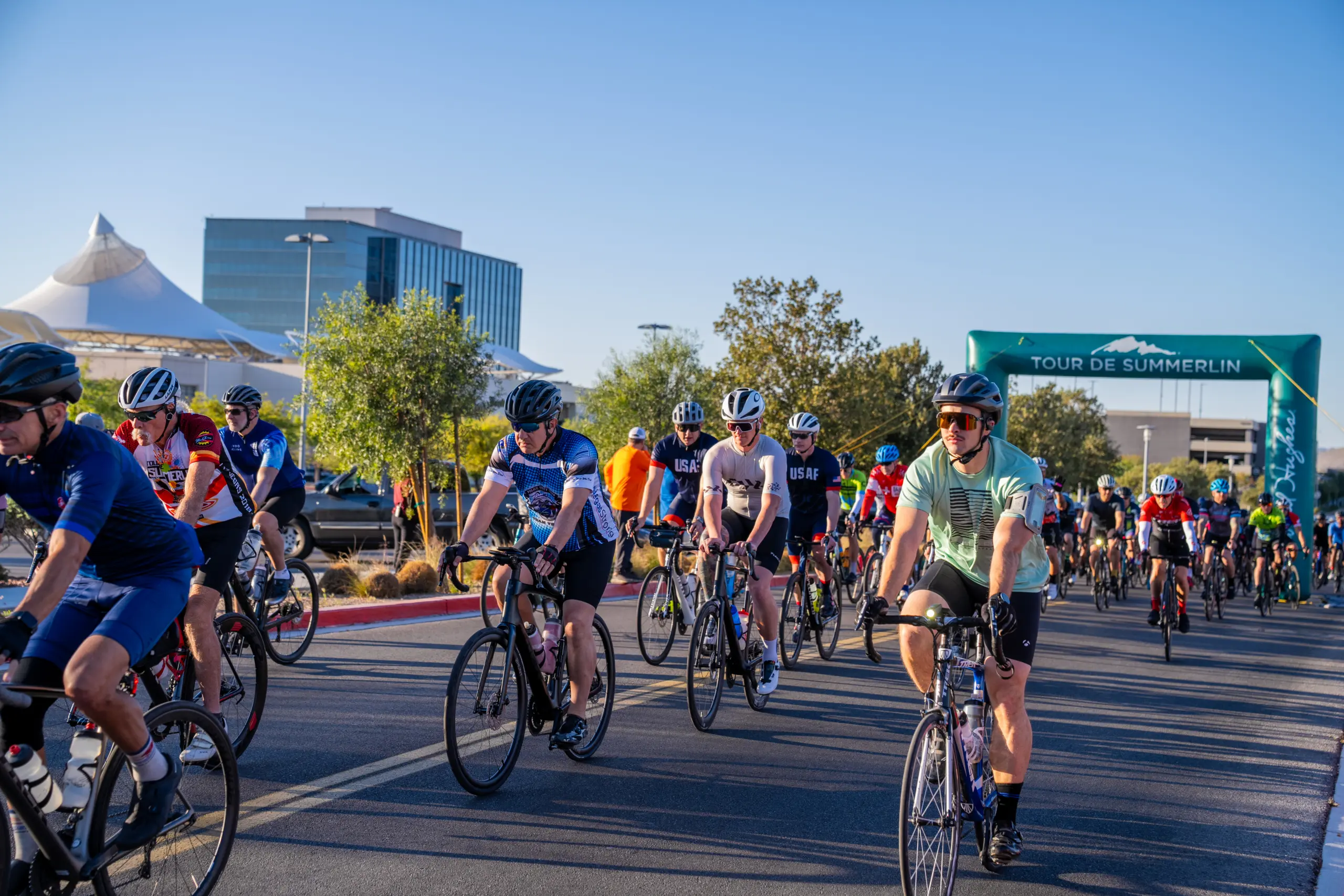 Cyclists participate in a group ride during the Tour de Summerlin event on a sunny day, surrounded by trees and modern buildings, under a clear blue sky.
