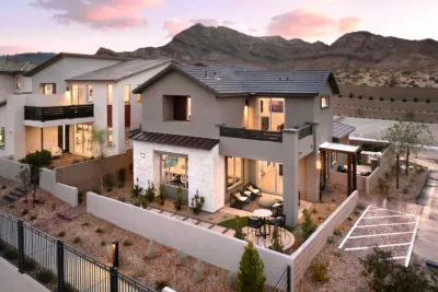 A modern two-story house with large windows, balconies, and a landscaped yard, set against a backdrop of mountains at sunset. The home features outdoor seating areas and a fenced yard with desert plants.