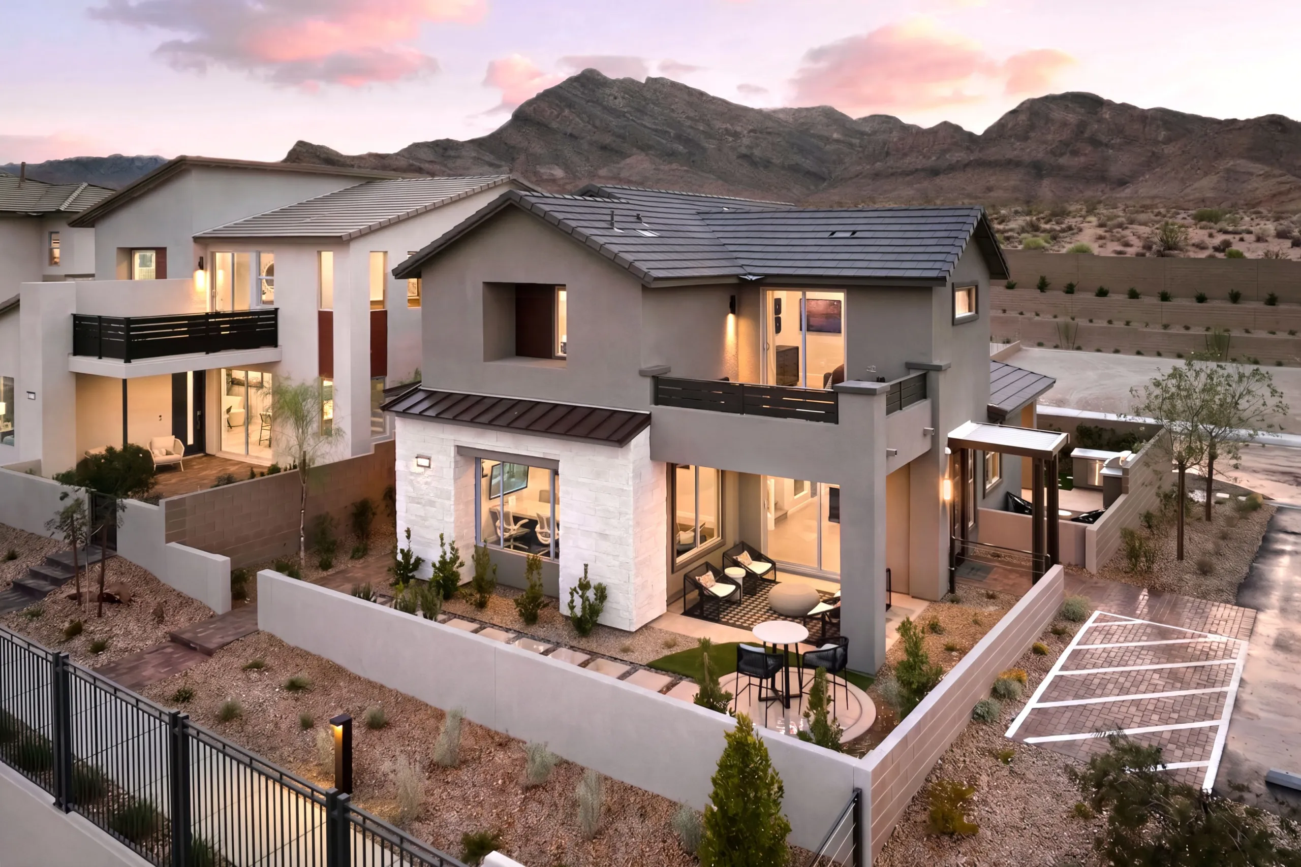 A modern two-story house with large windows, balconies, and a landscaped yard, set against a backdrop of mountains at sunset. The home features outdoor seating areas and a fenced yard with desert plants.