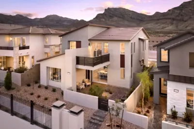Modern two-story house with large windows, a balcony, and a small courtyard, set in a desert landscape with rocky mountains in the background at sunset.