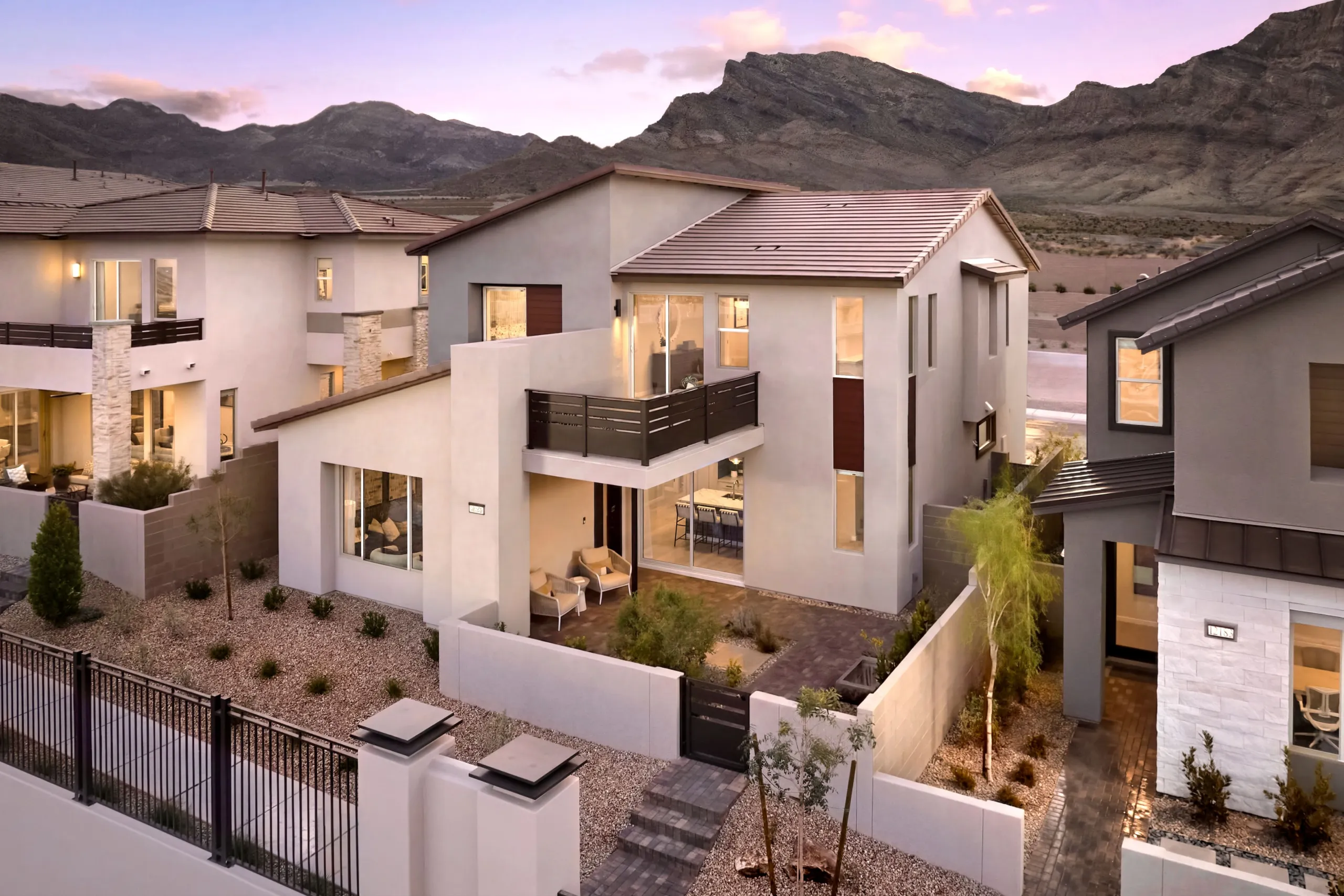 Modern two-story house with large windows, a balcony, and a small courtyard, set in a desert landscape with rocky mountains in the background at sunset.