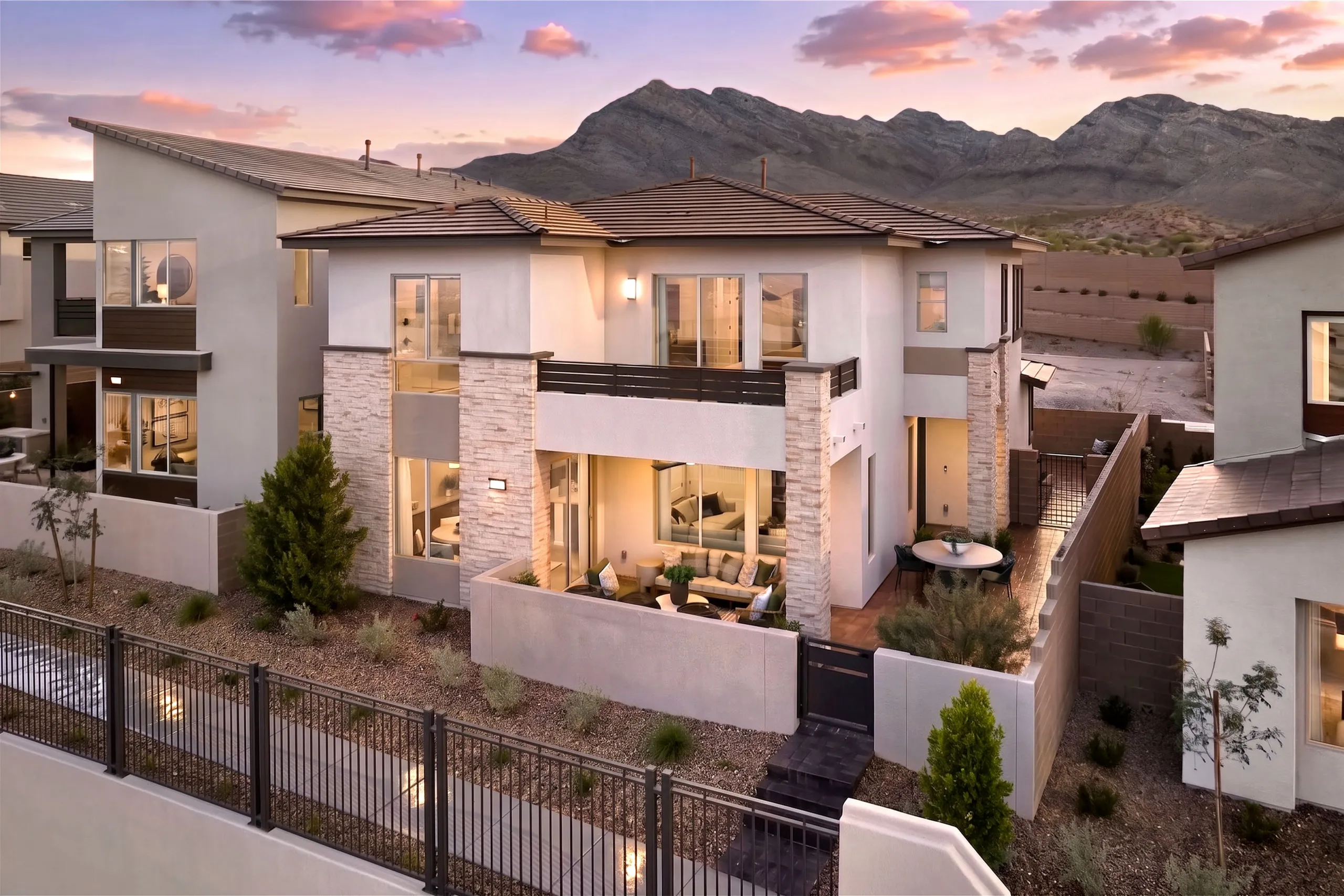 collection 1 plan 3 Modern two-story house with large windows and stone accents, outdoor seating areas, fenced yard, and mountain views in the background at sunset.